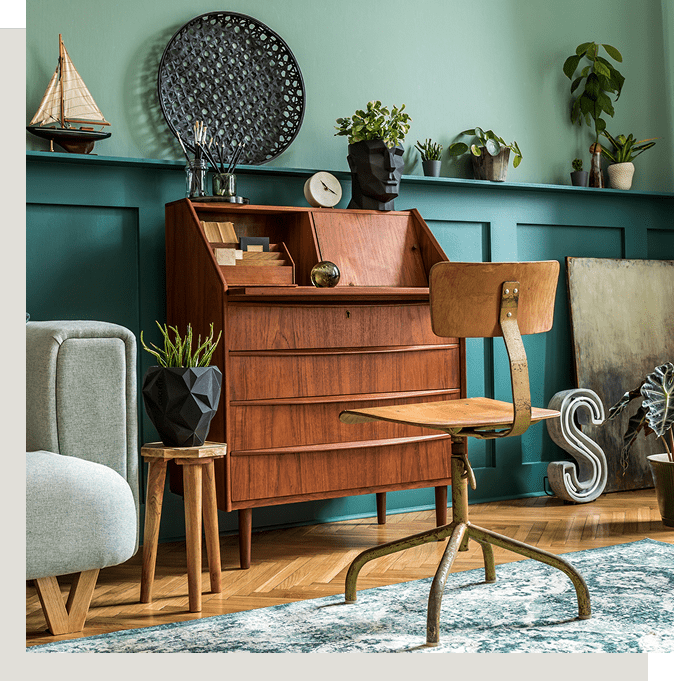 A mid-century modern room with a wooden desk, chair, and various plants. Decorative items include a ship model, round tray, and sculpted head. Pale green walls and parquet flooring complete the space.