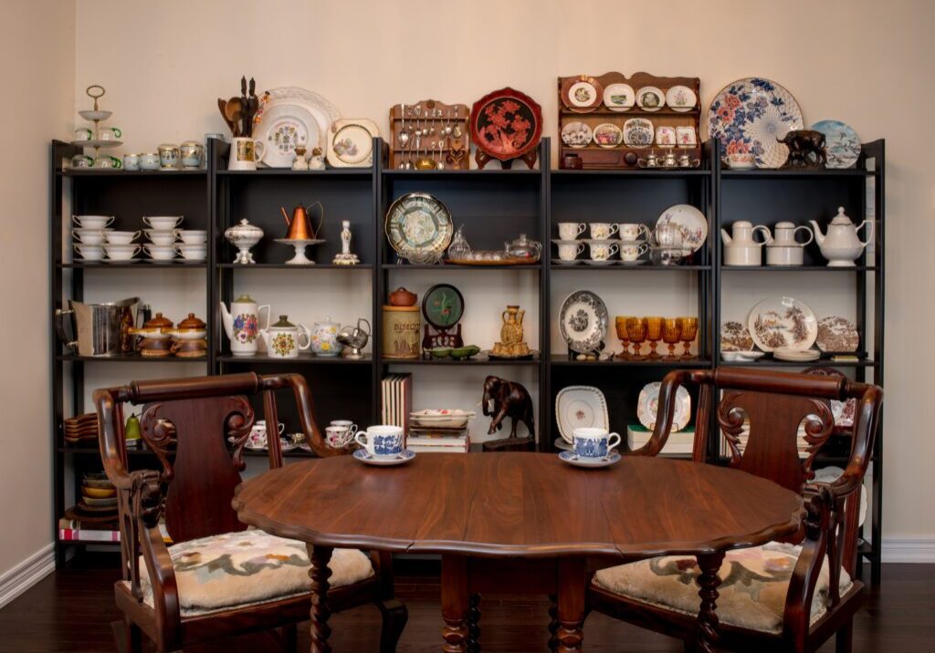 A room with a wooden table and chairs in the foreground. Background features shelves displaying various china plates, mugs, and decorative items.